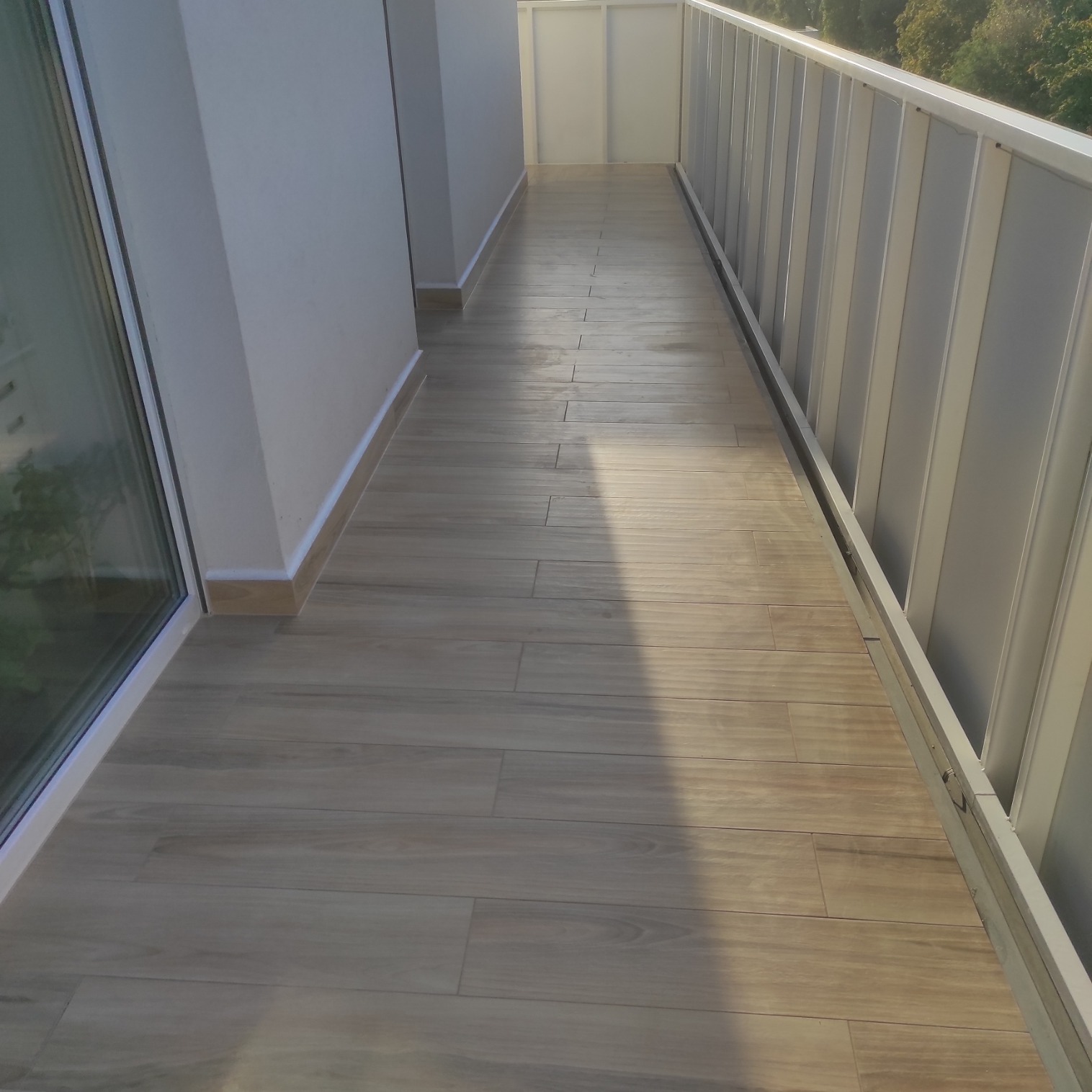 Wąski balkon wyłożony płytkami imitującymi drewno, z nowoczesną, białą balustradą i widokiem na zieleń za oknem.