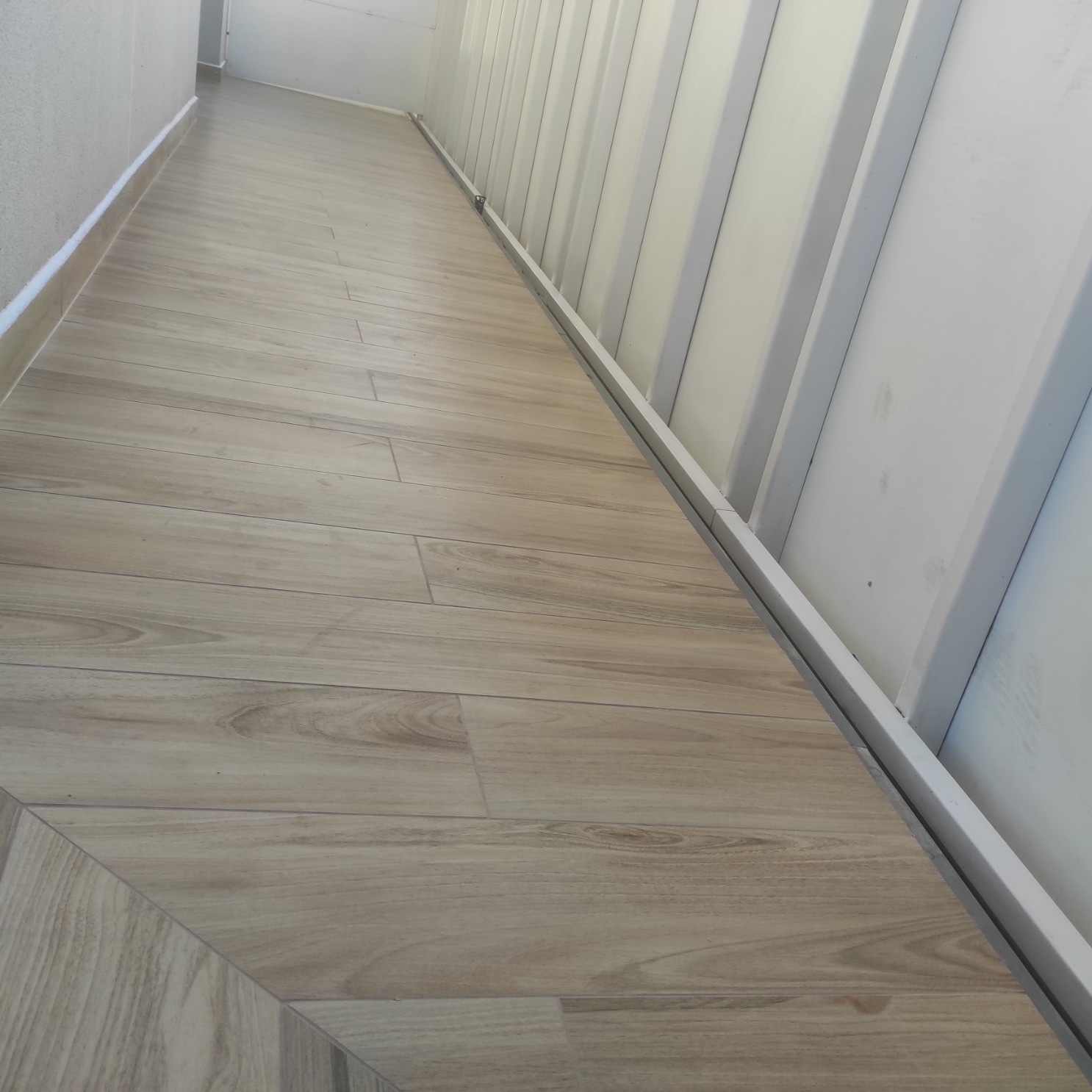 Ułożona pod kątem podłoga z płytek imitujących drewno na zabudowanym balkonie, widok z perspektywy podłogi w kierunku ściany z białymi panelami.