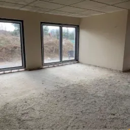 Surowy stan wnętrza z dużymi oknami przesuwnymi przed nałożeniem tynków, widoczna surowa podłoga i ściany przygotowane do prac wykończeniowych.