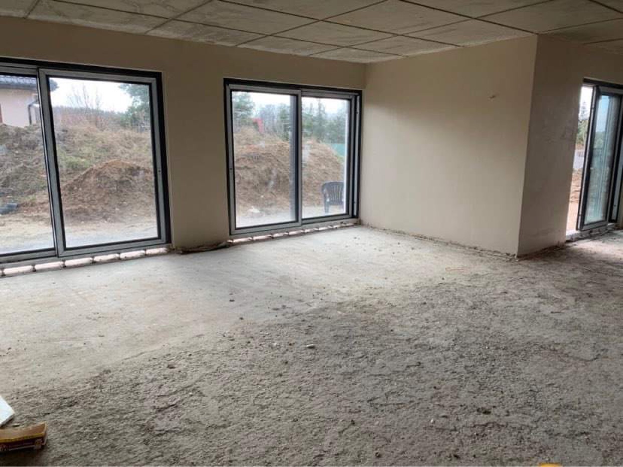 Surowy stan wnętrza z dużymi oknami przesuwnymi przed nałożeniem tynków, widoczna surowa podłoga i ściany przygotowane do prac wykończeniowych.