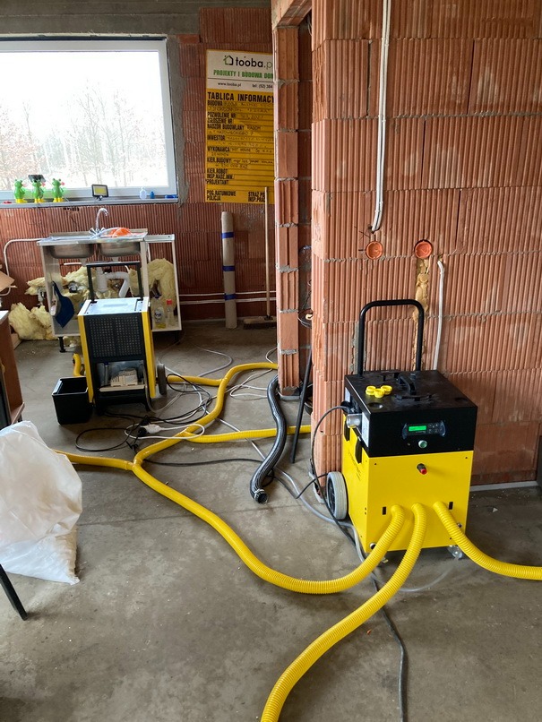 Dwa żółte urządzenia z podłączonymi elastycznymi przewodami na surowym betonowym podłożu w pomieszczeniu z ceglanymi ścianami w trakcie budowy; w tle zlew na metalowym stelażu.