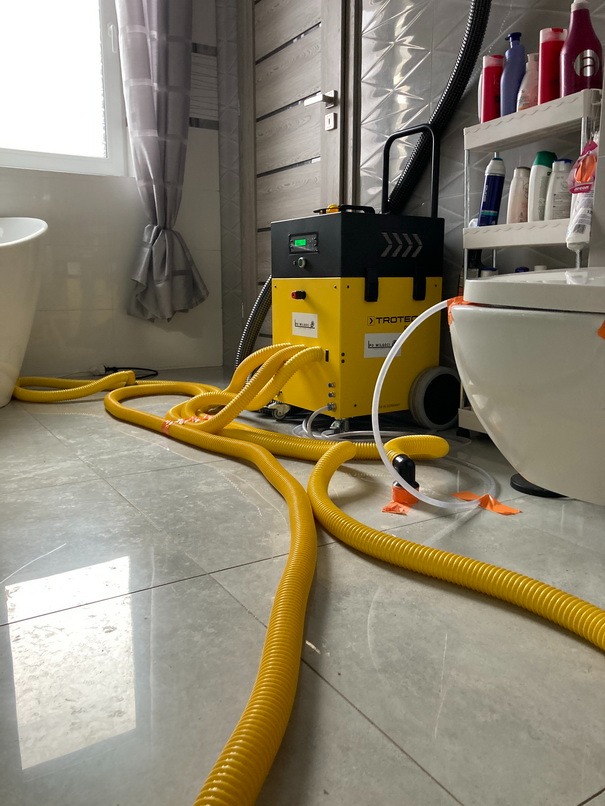 Żółty osuszacz kondensacyjny Trotec z wieloma wężami w łazience z szarymi płytkami, widoczne okno i wanna