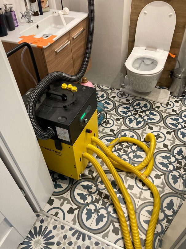 Żółty profesjonalny osuszacz Trotec z podłączonymi elastycznymi wężami w łazience z wzorzystą podłogą, widoczna umywalka i toaleta.