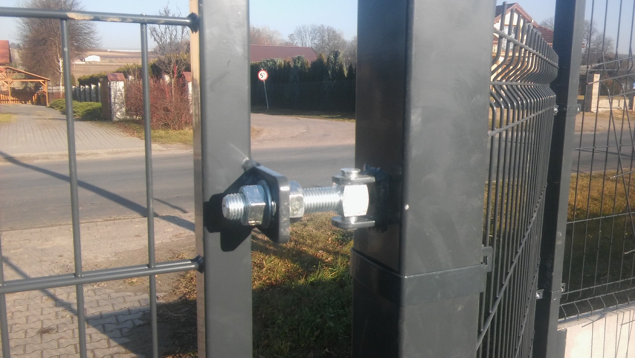 Szczegół mocowania bramy panelowej, widoczne śruby i nakrętki łączące metalowe słupki, w tle ulica i zabudowa jednorodzinna.
