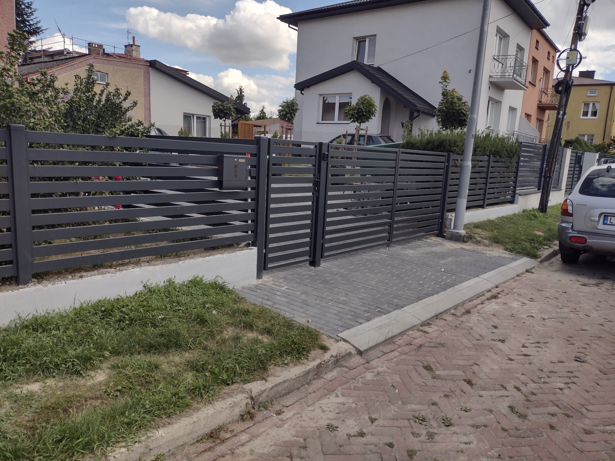 Nowoczesne, poziome przęsła w kolorze grafitowym tworzą ogrodzenie posesji z bramą wjazdową, uzupełnione skrzynką na listy, na tle zabudowy jednorodzinnej.