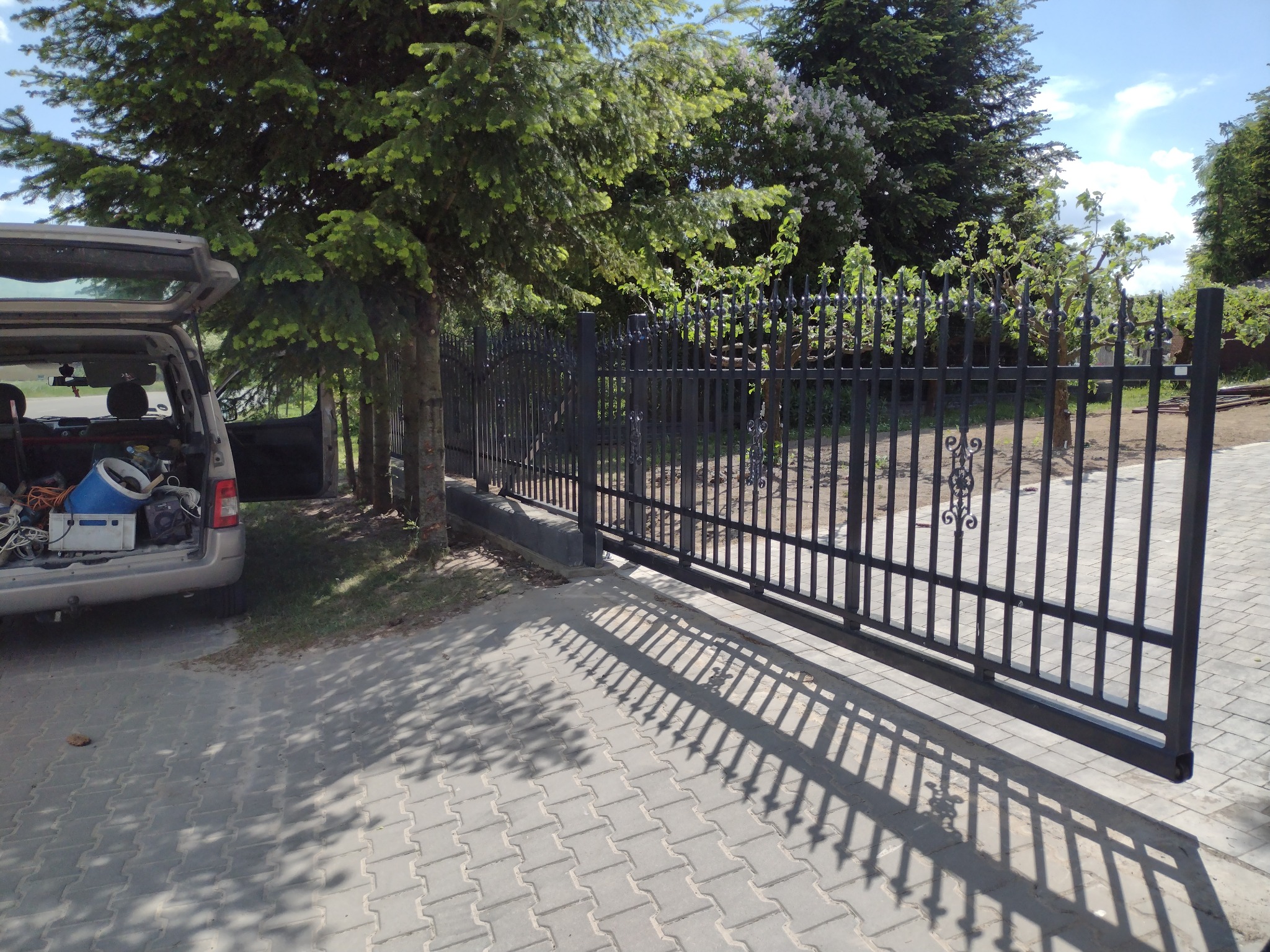 Zasuwna, metalowa brama przesuwna w kolorze czarnym, z widocznymi cieniami na szarej kostce brukowej, samochód z otwartym bagażnikiem i narzędziami w tle.