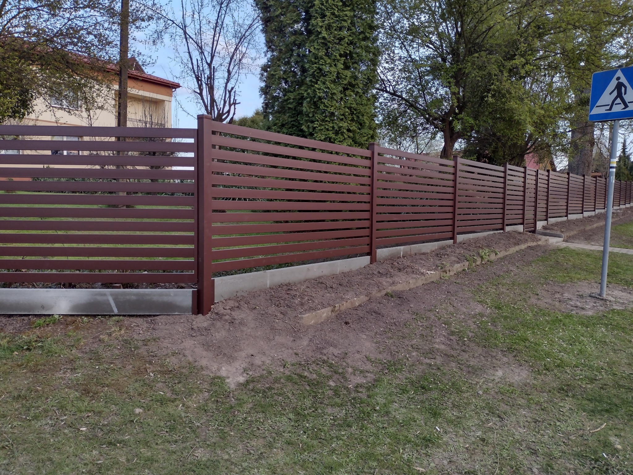 Długi, poziomy, brązowy płot z metalowych paneli z betonową podmurówką wzdłuż trawnika, z widocznym znakiem przejścia dla pieszych w tle.
