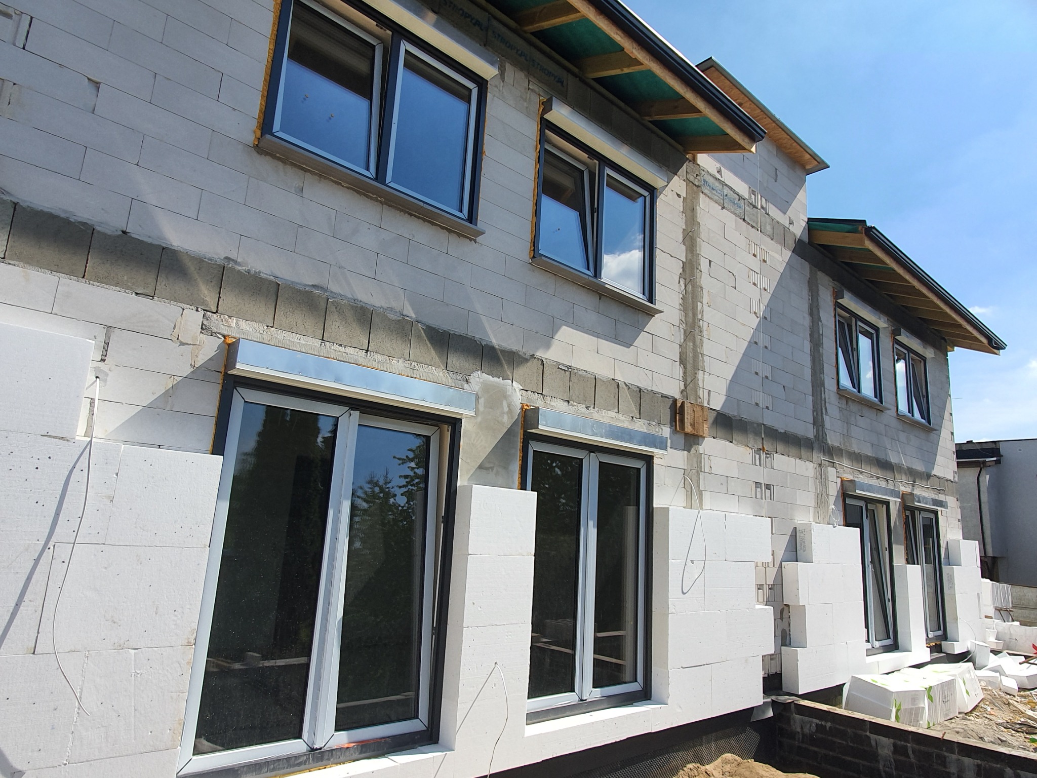 Niedokończony budynek z białych bloczków, z zamontowanymi oknami z czarnymi ramami, ocieplony białym styropianem, widok z dołu na fragment elewacji i dachu.