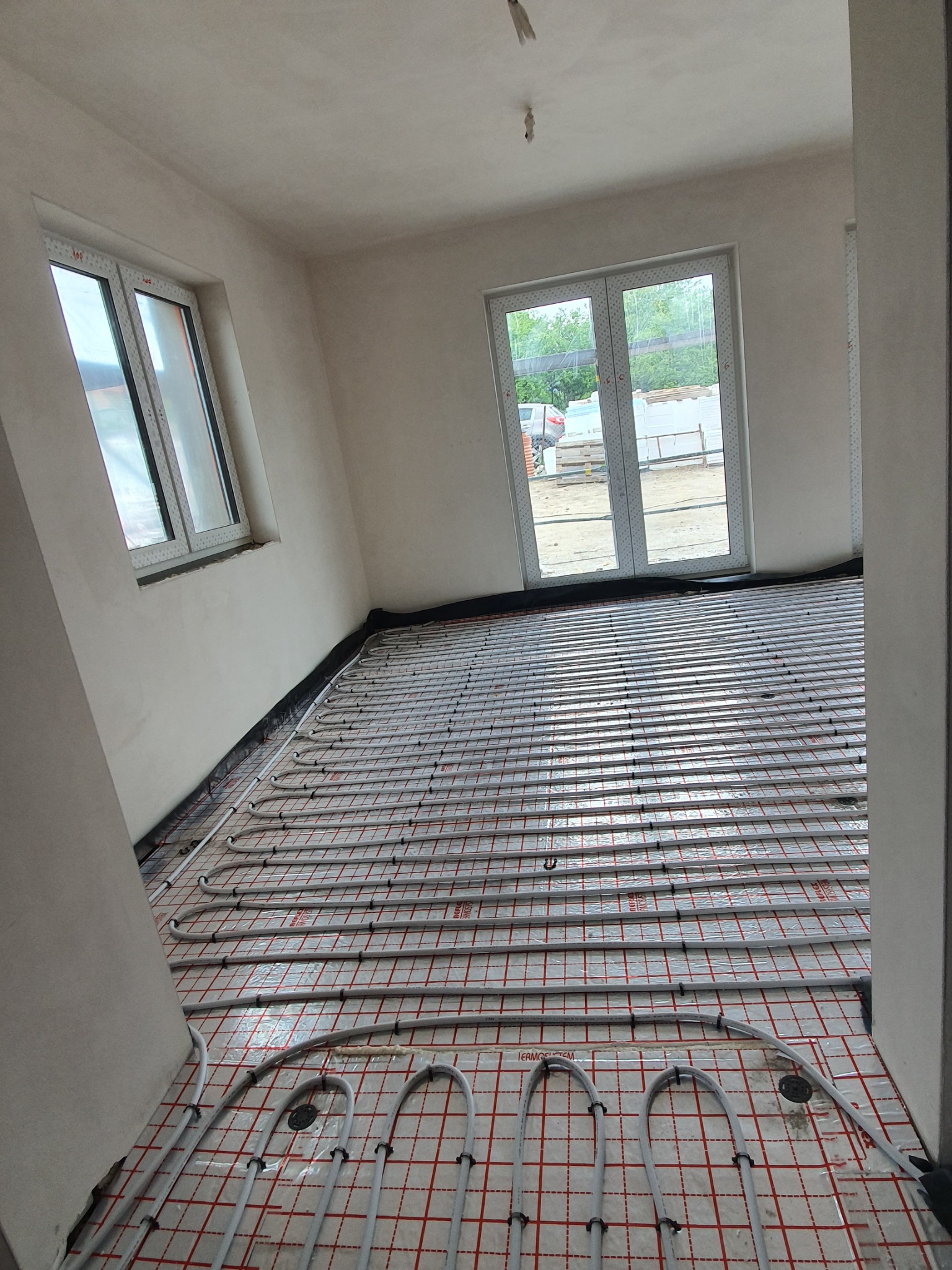 Wnętrze w stanie surowym z instalacją ogrzewania podłogowego rozłożoną na folii z czerwoną kratką; widoczne okna i drzwi balkonowe.