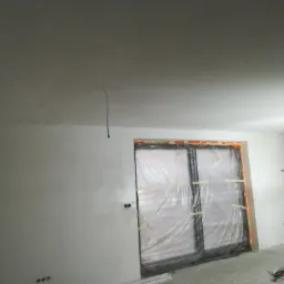 Świeżo wyszpachlowana ściana z widocznymi punktami elektrycznymi, zabezpieczone okno folią, widoczny fragment drabiny i wałka malarskiego, przygotowane narzędzia na podłodze.