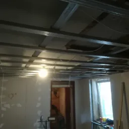 Montaż metalowej konstrukcji sufitu podwieszanego z widocznymi przewodami elektrycznymi i oświetleniem tymczasowym, ściany częściowo pokryte płytami gipsowo-kartonowymi z widocznymi miejscami...