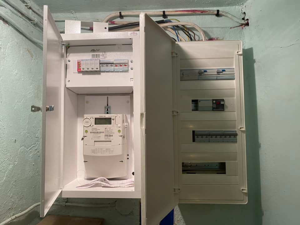 Otwarta skrzynka elektryczna z licznikiem i zabezpieczeniami, widoczne kable w ścianie, surowy stan wykończenia wnętrza.