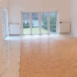 Przygotowanie podłoża pod nową podłogę w jasnym, przestronnym pokoju z marmurową posadzką i widokiem na ogród przez duże okna.