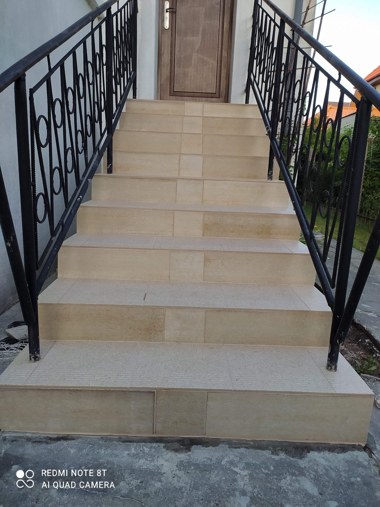 Wyłożone jasnobeżowymi płytkami schody wejściowe z czarną metalową balustradą, prowadzące do brązowych drzwi wejściowych.