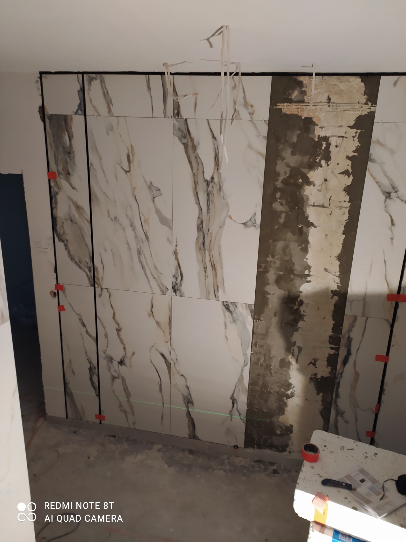 Montaż płytek imitujących marmur na ścianie, widoczne krzyżyki dystansowe i fragment ściany z nałożonym klejem, laserowy niwelator wyznacza linię.