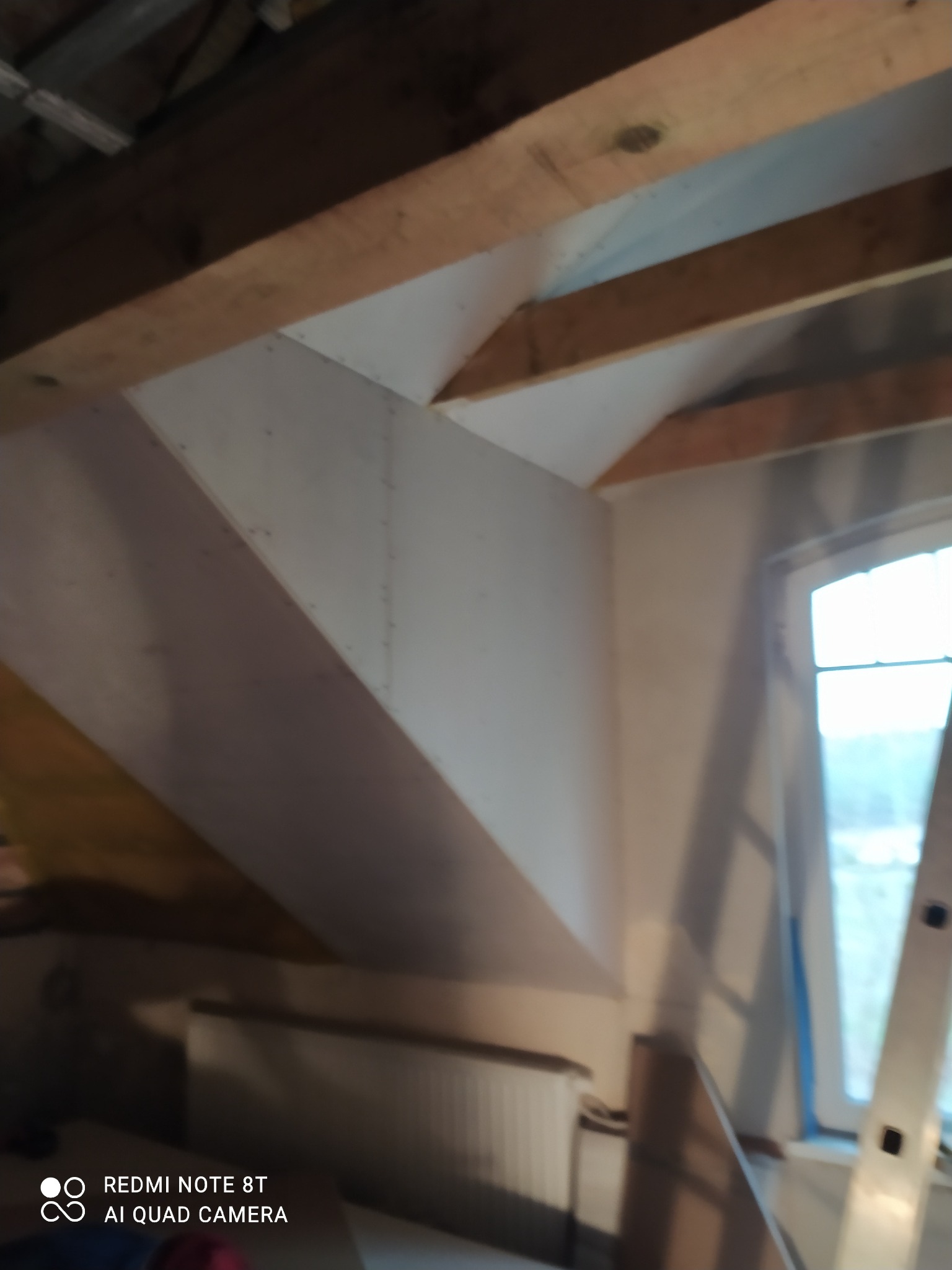 Poddasze w trakcie prac wykończeniowych: drewniane belki konstrukcyjne, ściany pokryte płytami gipsowo-kartonowymi, widoczne okno i grzejnik, zdjęcie rozmazane.