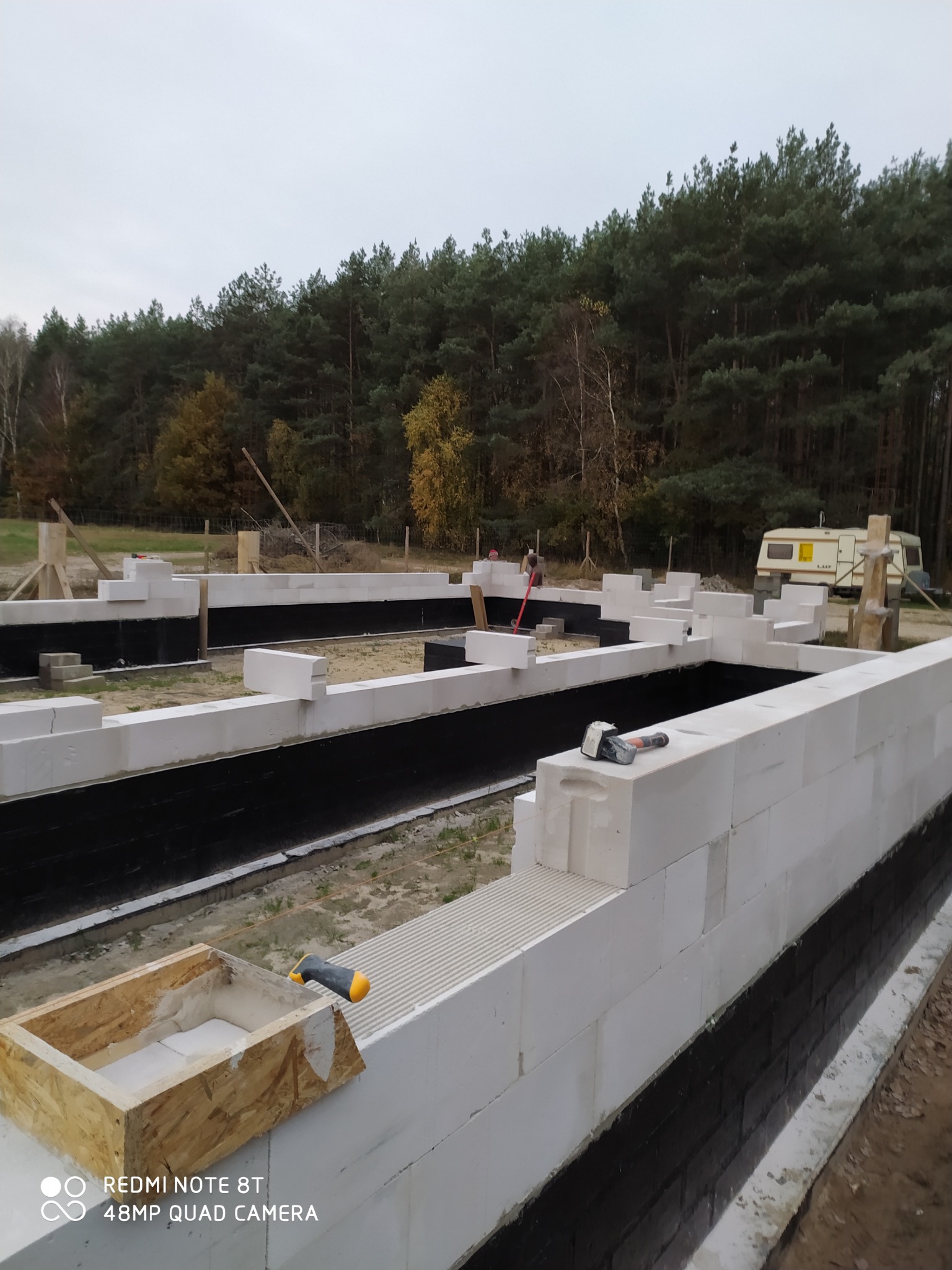 Budowa fundamentów domu z bloczków betonowych, zabezpieczonych czarną izolacją, widok na las w tle.