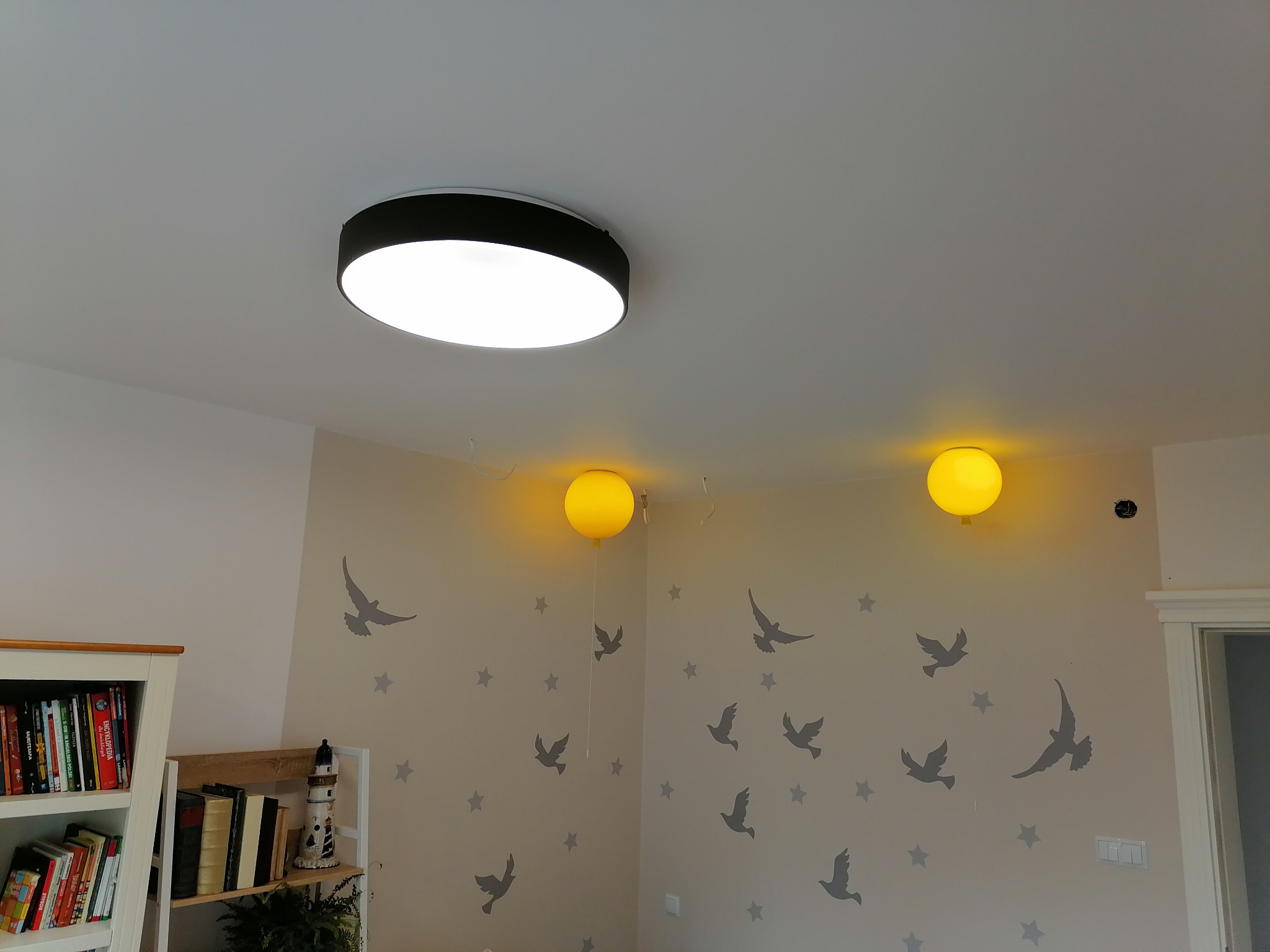 Pokój dziecięcy z trzema lampami: okrągłą sufitową i dwoma ściennymi w kształcie żółtych balonów, ściany zdobione naklejkami gwiazd i ptaków.