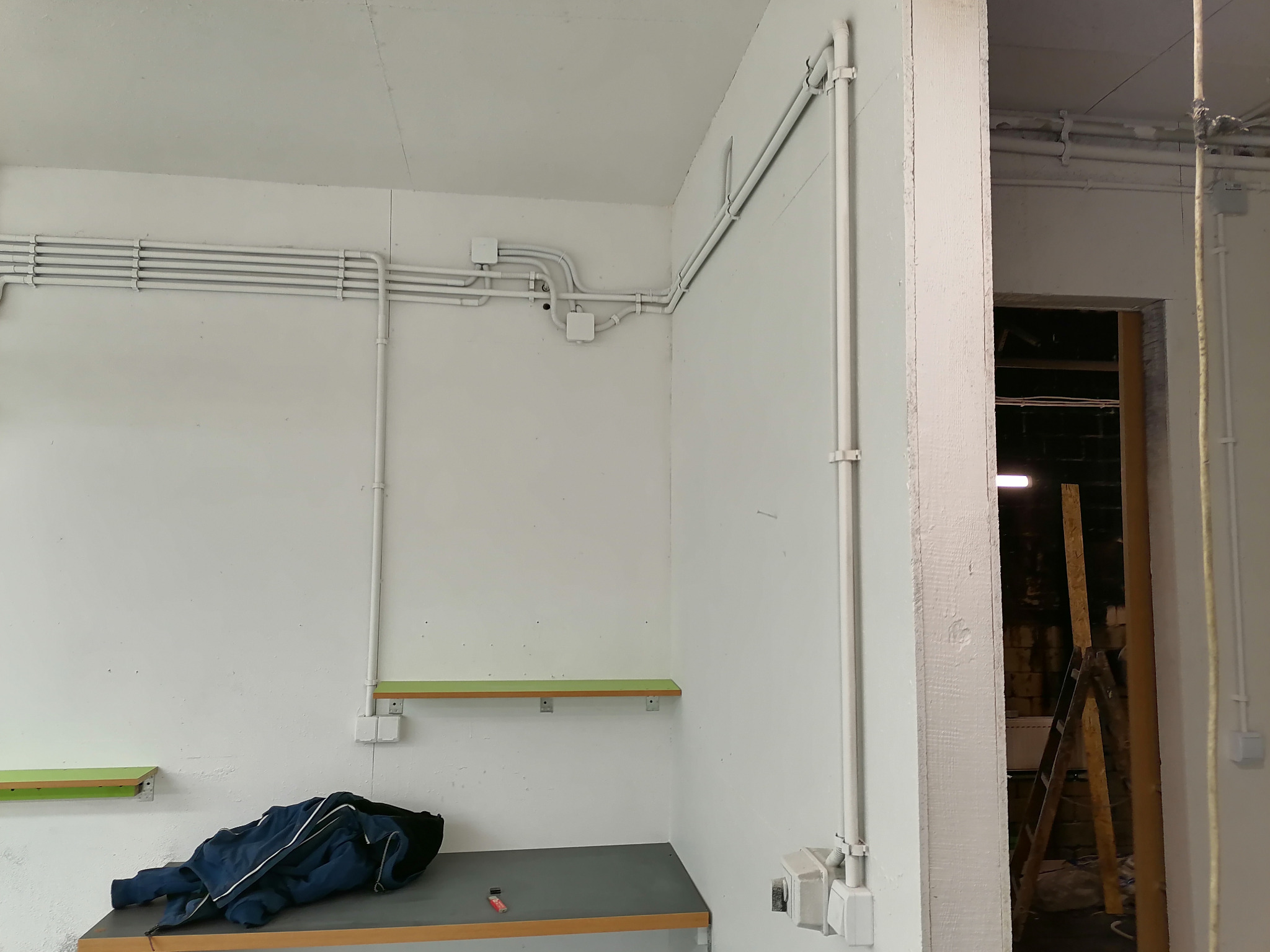 Białe rury instalacji elektrycznej poprowadzone po ścianach w pomieszczeniu z surowym wykończeniem, widoczne gniazdka, półki z zielonymi krawędziami oraz ubranie na blacie, w tle otwarte przejście...