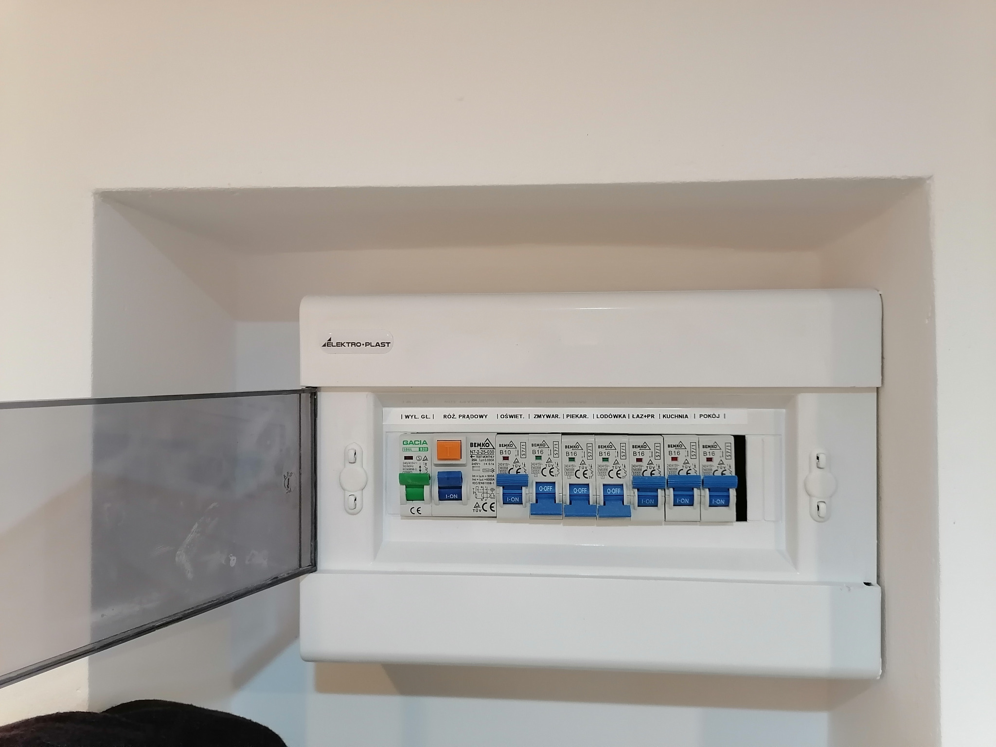 Rozdzielnica elektryczna marki Elektro-Plast, zamontowana we wnęce ściany, z uchylonymi drzwiczkami po lewej stronie, widoczne bezpieczniki z opisami obwodów.