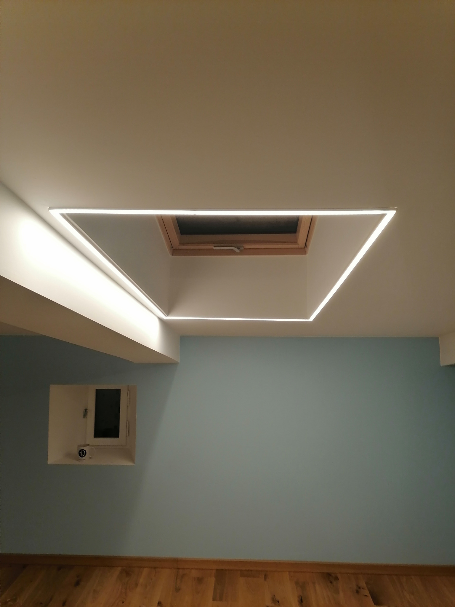 Wnętrze pokoju z oknem dachowym otoczonym kwadratową ramą z oświetleniem LED, ściany w odcieniach błękitu, drewniana podłoga i małe okienko w ścianie z kubkiem na parapecie.