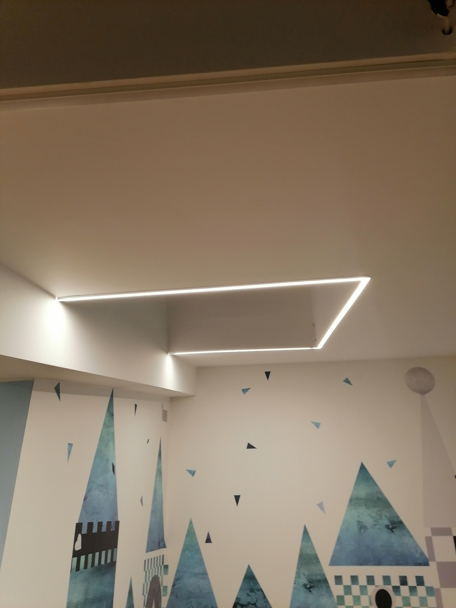 Wpuszczone w sufit prostokątne oświetlenie LED w pokoju dziecięcym z malowanymi na ścianie elementami zamku i trójkątami.