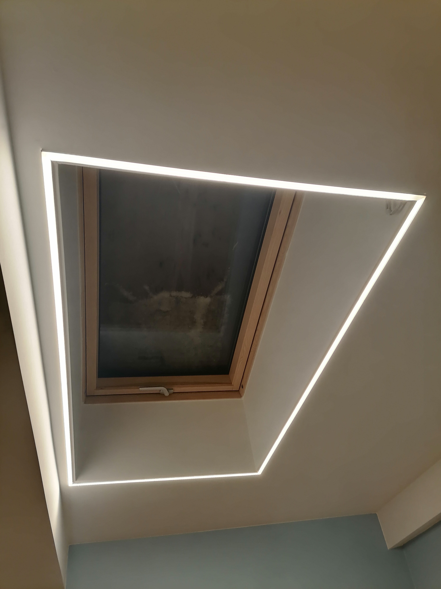 Oświetlenie LED, tworzące geometryczną ramę wokół okna dachowego, w minimalistycznym wnętrzu z jasnymi ścianami.