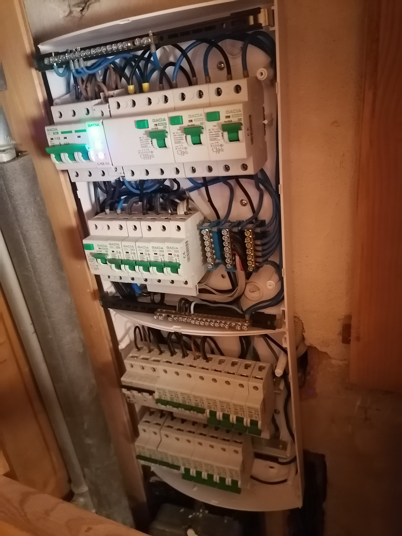 Otwarta skrzynka rozdzielcza z zamontowanymi bezpiecznikami i podłączonymi przewodami w kolorze niebieskim i czarnym, widoczne listwy zaciskowe i elementy montażowe, obok ściana z widocznym...