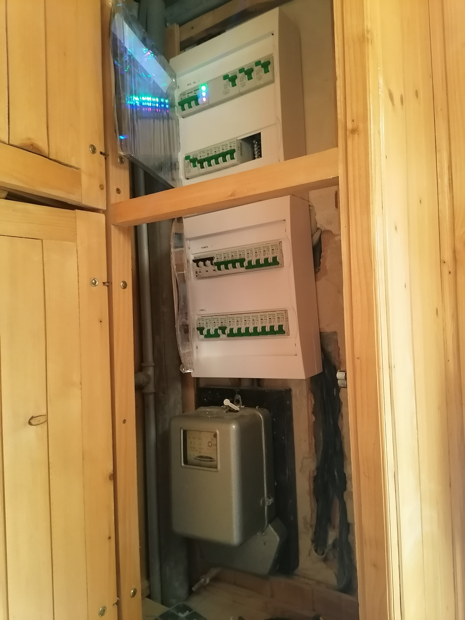 Dwa białe rozdzielnice elektryczne zamontowane na ścianie w drewnianej obudowie, widoczny również licznik elektryczny i fragment instalacji.