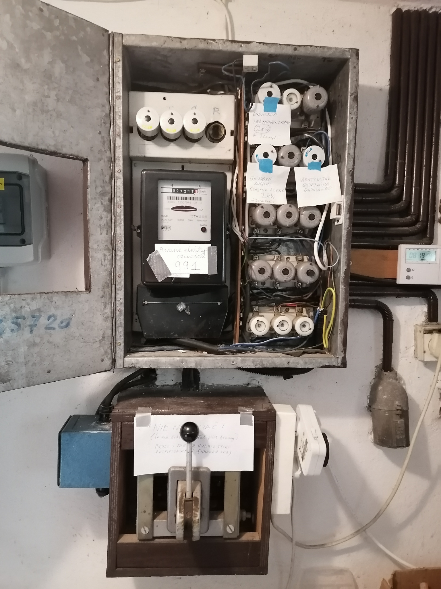 Otwarta, metalowa skrzynka z licznikiem energii elektrycznej, bezpiecznikami topikowymi i wyłącznikiem głównym. Widoczne notatki przyklejone do elementów instalacji.