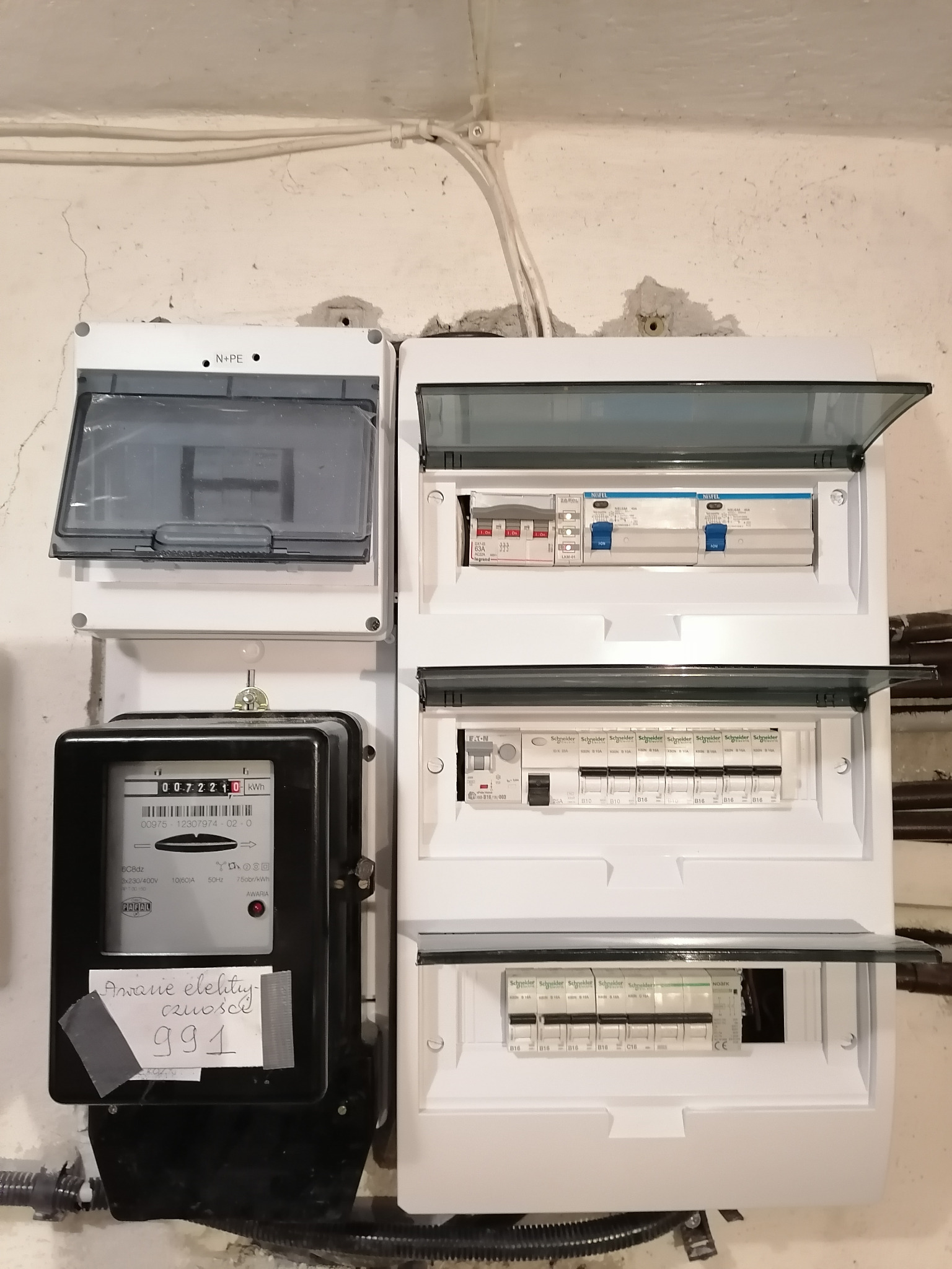 Rozdzielnica elektryczna z licznikiem, widoczne zabezpieczenia różnicowoprądowe i nadprądowe, kartka z napisem 'Awarie elektryczne czułość 991' przyklejona do licznika.