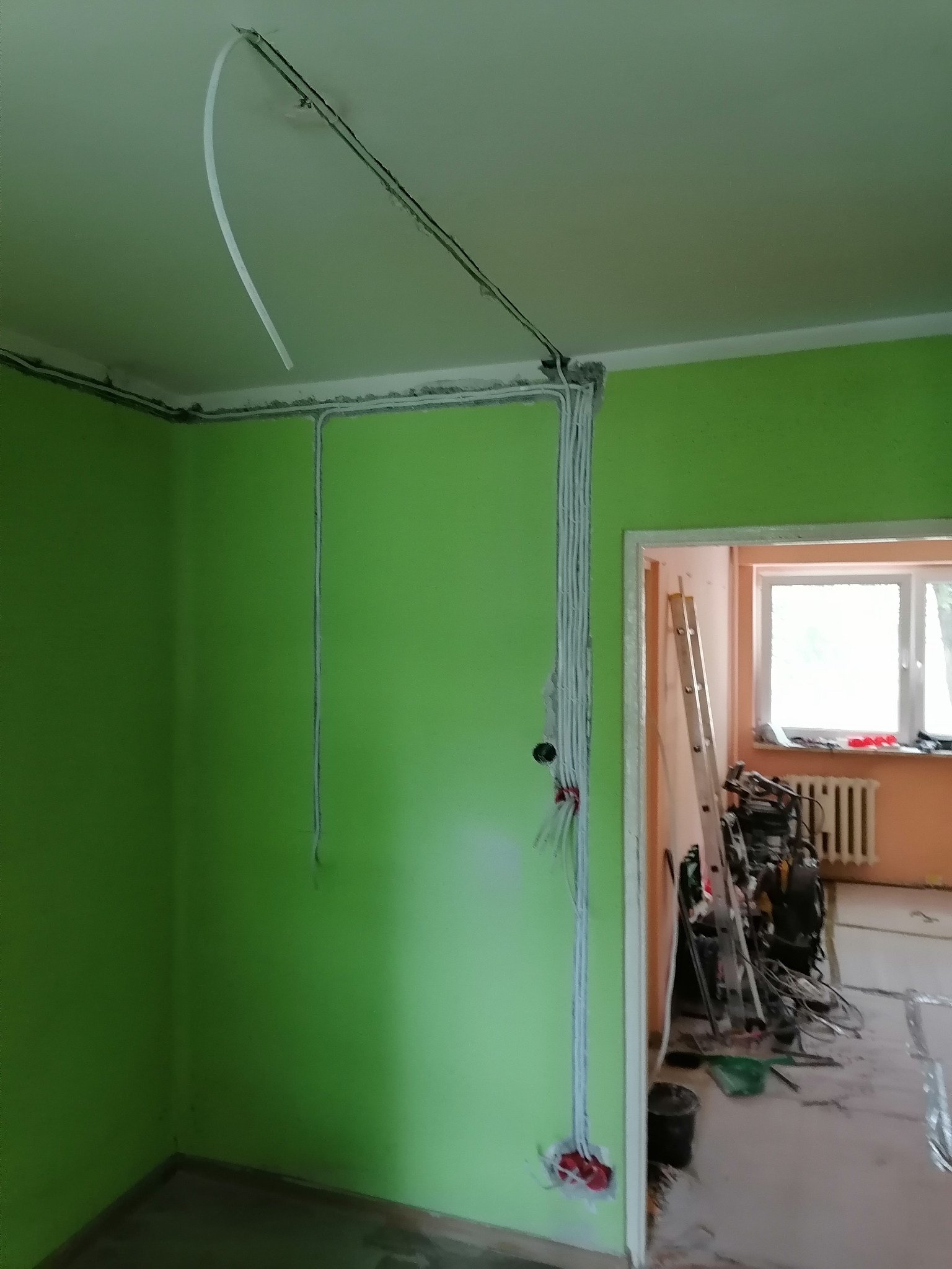 Świeżo pomalowane na zielono wnętrze pokoju z widocznymi przewodami elektrycznymi prowadzonymi w listwach przypodłogowych i pod sufitem, z puszkami elektrycznymi w ścianie i przewodami wystającymi...