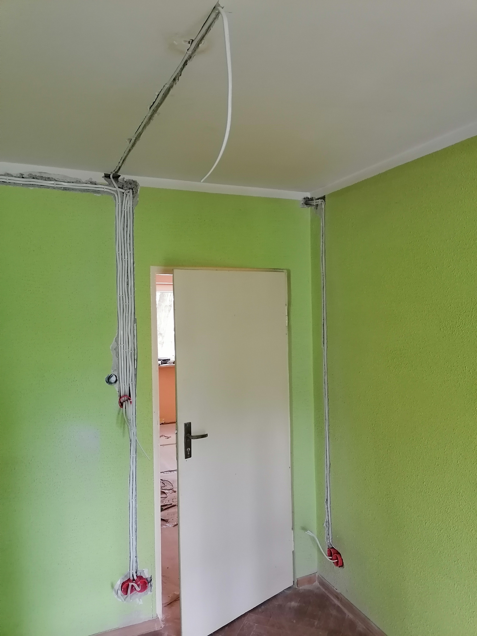 Wnętrze pokoju z zielonymi ścianami w trakcie montażu instalacji elektrycznej: kable prowadzone w bruzdach, częściowo zamaskowane gipsem, widoczne puszki instalacyjne z wystającymi przewodami.