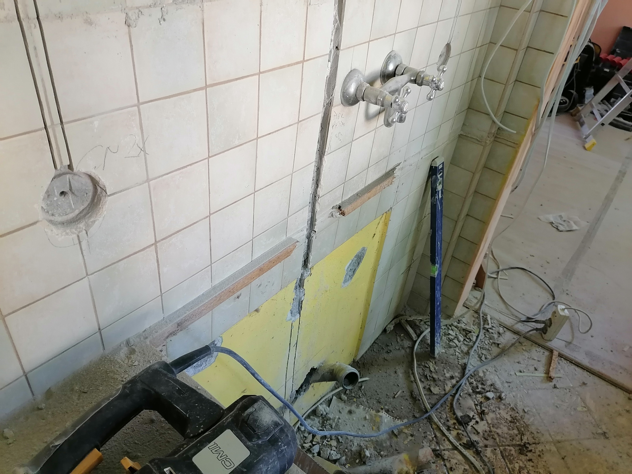 Remont łazienki: usunięte płytki, odsłonięte instalacje elektryczne i hydrauliczne, widoczny przewód elektryczny wychodzący ze ściany i kable na podłodze, narzędzie do kucia płytek