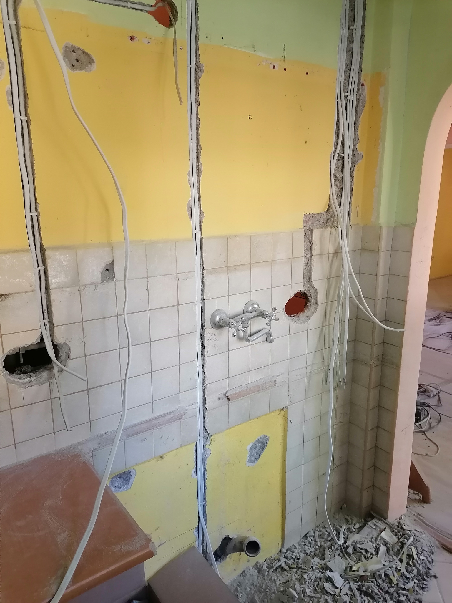 Ściana w trakcie remontu z widocznymi przewodami elektrycznymi w bruzdach, fragmentem starej armatury i pozostałościami po skuciu płytek.