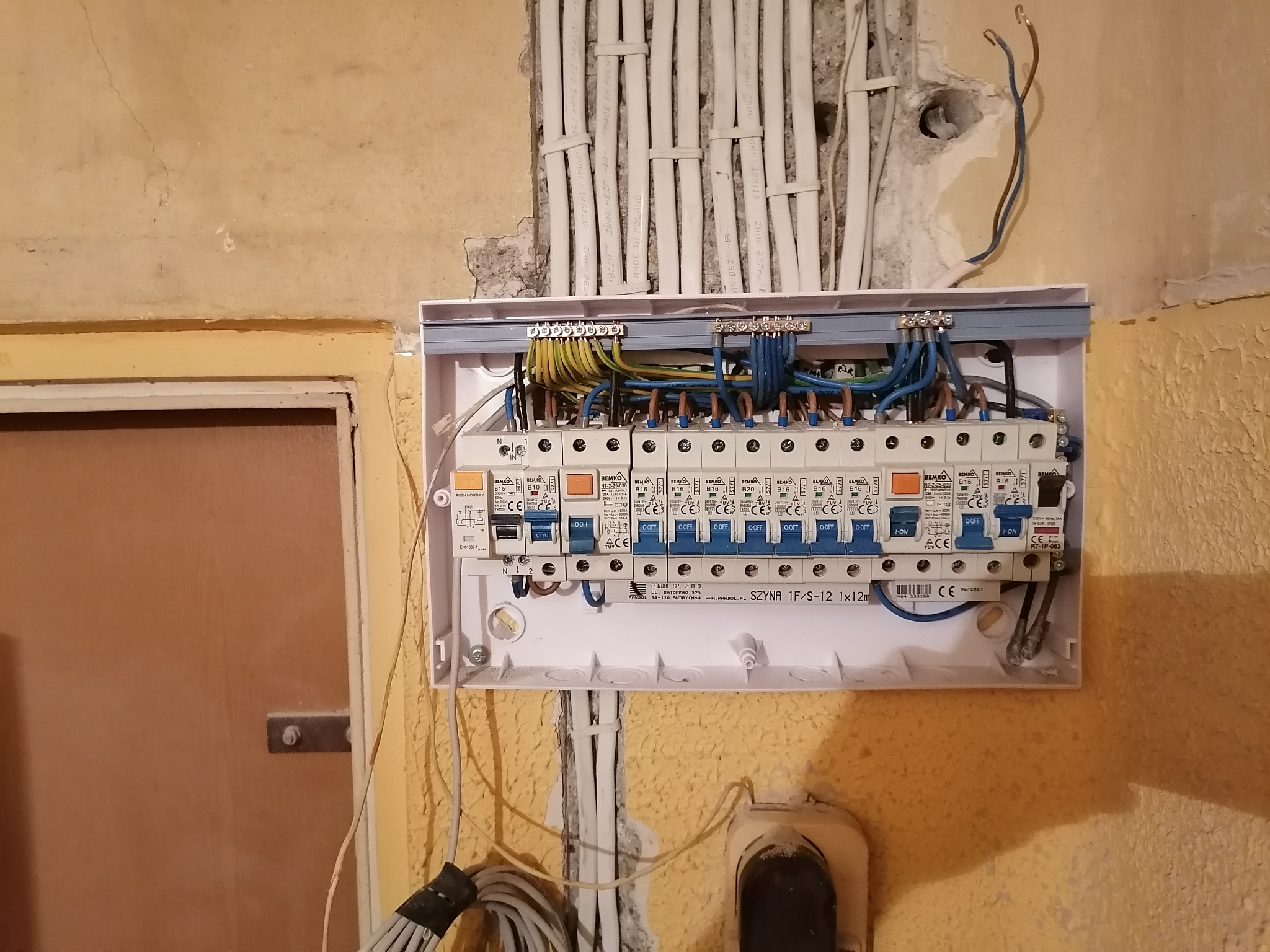 Otwarta skrzynka rozdzielcza z zamontowanymi bezpiecznikami i podłączonymi przewodami elektrycznymi na nierównej, żółtej ścianie. Widoczne białe kable w ścianie oraz fragment drzwi po lewej stronie.
