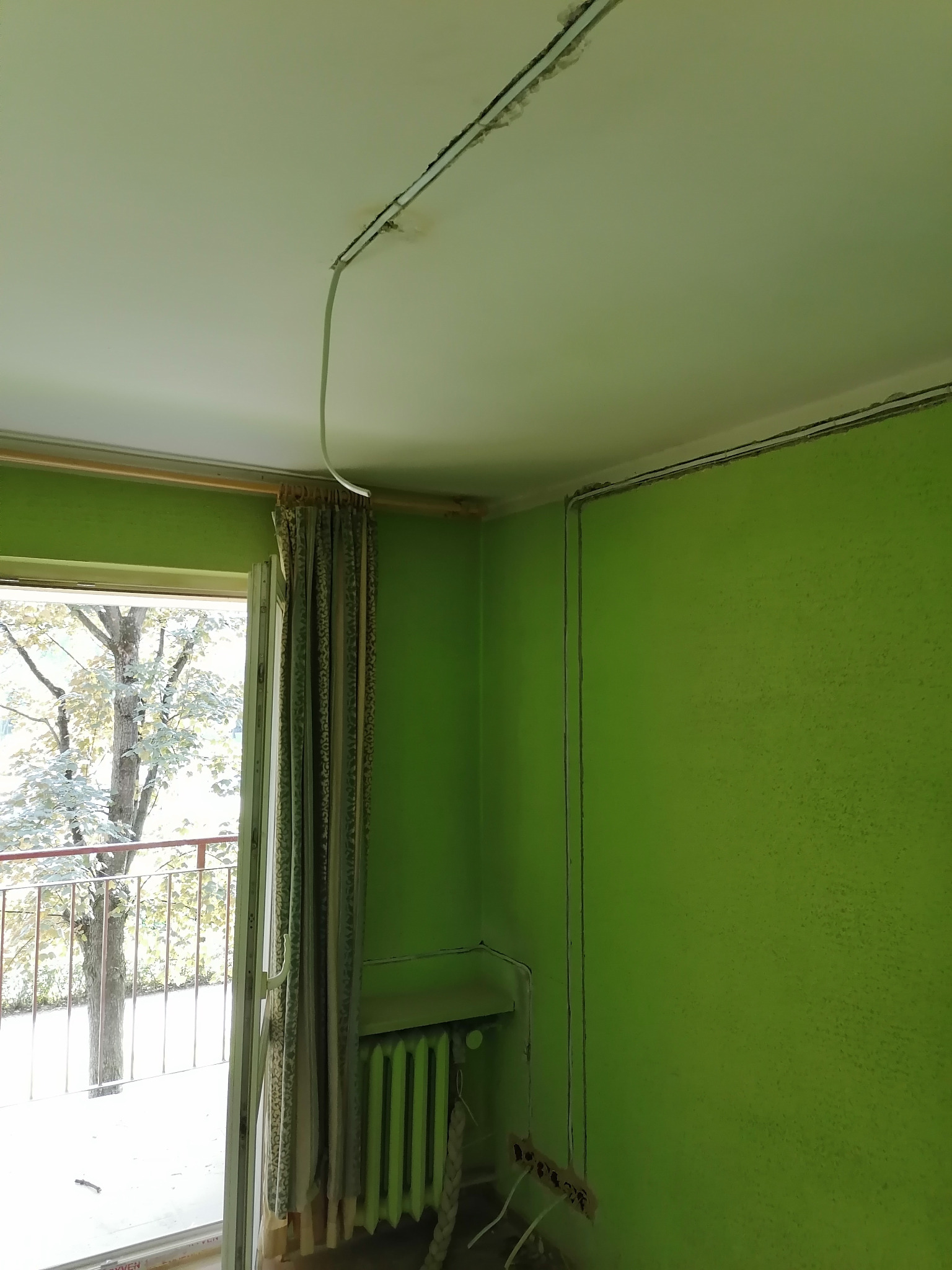 Pokój z zielonymi ścianami w trakcie remontu, widoczne kable elektryczne prowadzone po suficie i ścianach, oraz wystające przewody z gniazdek. Grzejnik pomalowany na zielono, okno balkonowe z zasłoną.