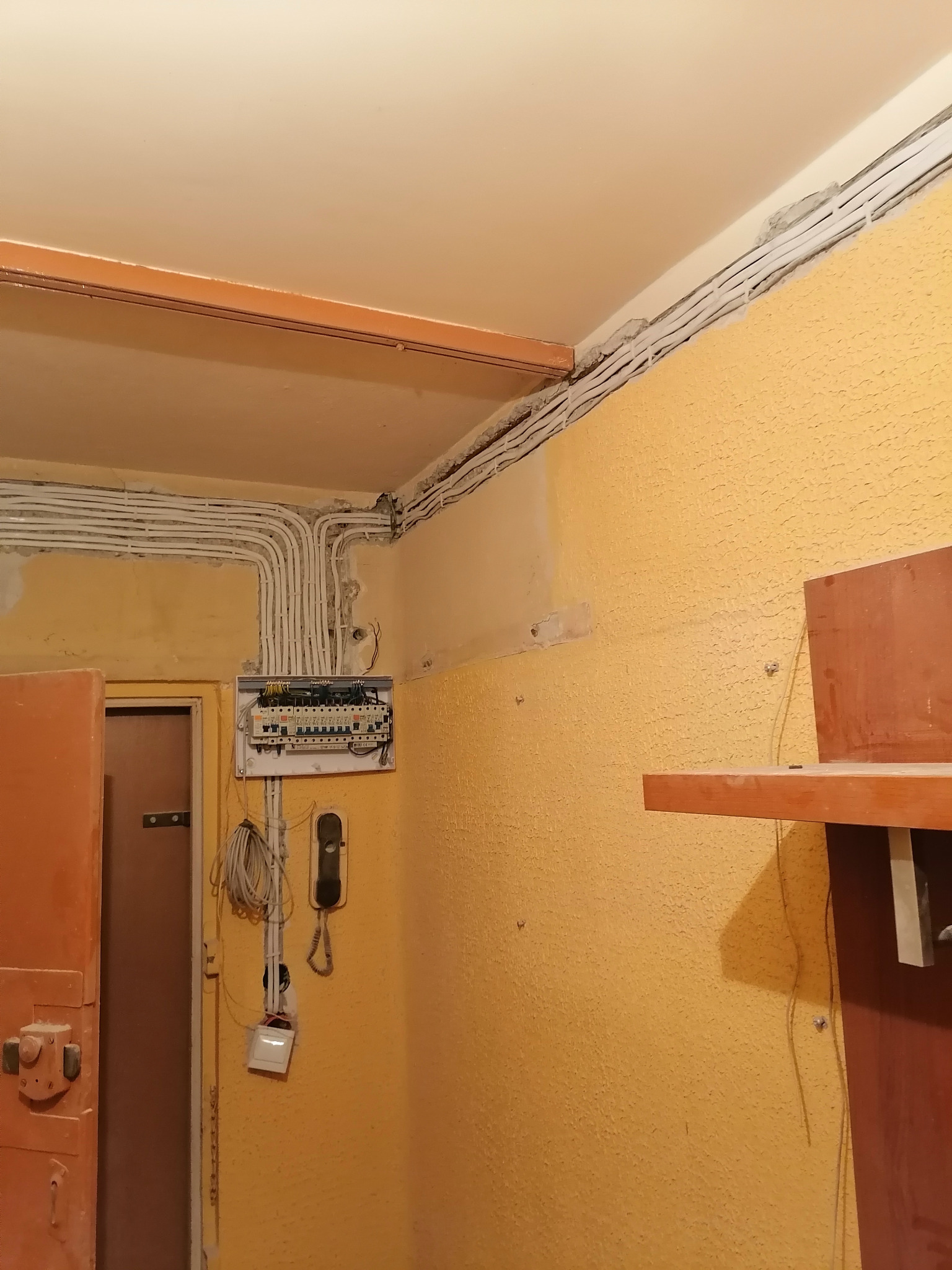 Kable elektryczne prowadzone po ścianie i suficie, z widoczną skrzynką bezpiecznikową w mieszkaniu o żółtych ścianach i pomarańczowych elementach wykończeniowych.