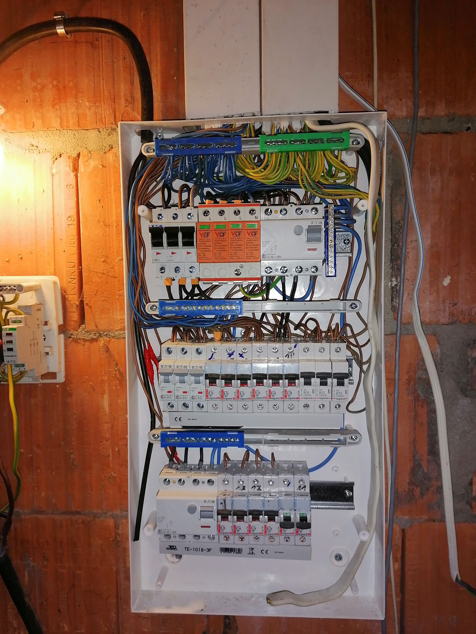 Rozdzielnica elektryczna z bezpiecznikami, przewodami i listwami zaciskowymi zamontowana na ceglanej ścianie. Widoczne różne typy zabezpieczeń i okablowania. Część przewodów wychodzi poza obudowę...