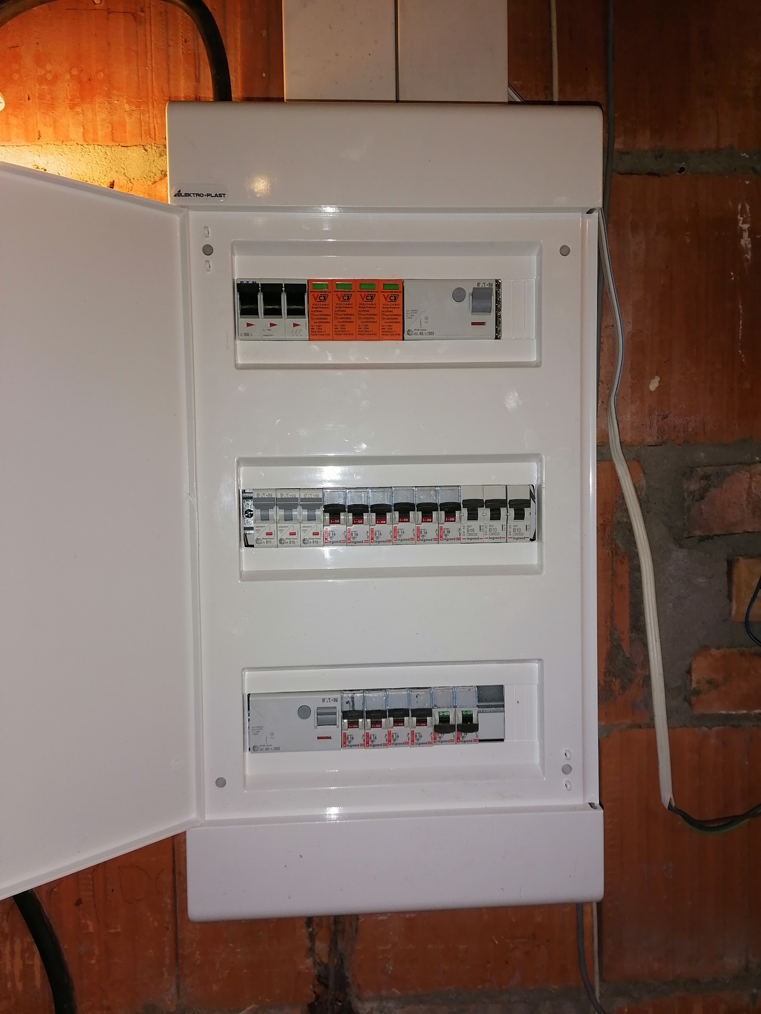 Otwarta, biała skrzynka rozdzielcza z bezpiecznikami na ścianie z czerwonej cegły, widoczne kable i logo Elektro-Plast.