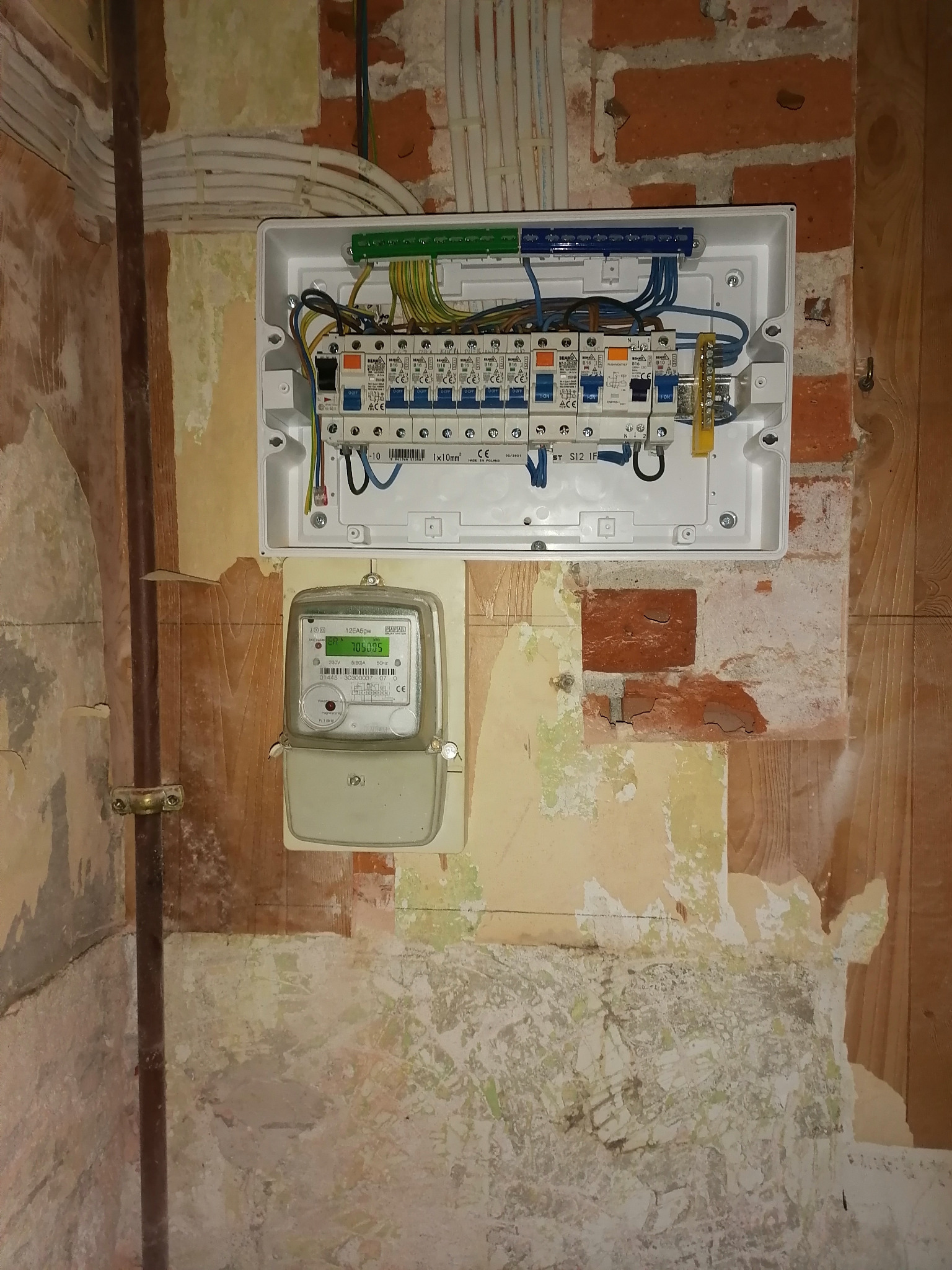 Rozdzielnica elektryczna natynkowa z zabezpieczeniami i przewodami, obok licznik energii elektrycznej na ścianie z widocznymi śladami po starych tapetach i odsłoniętą cegłą.