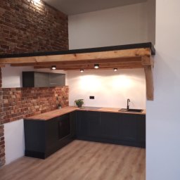 Łukasz Maryniak - Nowoczesna kuchnia w Świętochłowicach z ciemnymi szafkami, drewnianym blatem i ścianą z cegły, oświetlona punktowymi lampami, z widocznym drewnianym stropem i jasną podłogą.