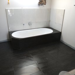 Łukasz Maryniak - Wnętrze łazienki w trakcie prac wykończeniowych; widoczna wanna obudowana ciemnymi płytkami, białe płytki na ścianie, ciemna podłoga z płytek oraz odkryte przewody elektryczne w ścianie.