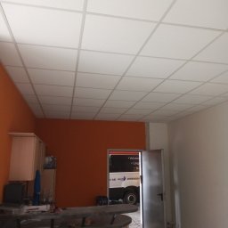 Łukasz Maryniak - Sufit podwieszany kasetonowy w pomieszczeniu z pomarańczową ścianą, widoczny fragment baru z krzesłami i otwarte drzwi z widokiem na samochód dostawczy.