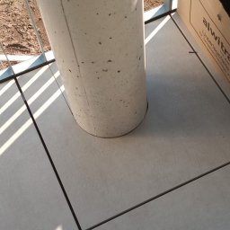 Łukasz Maryniak - Szare płytki podłogowe na balkonie z betonowym słupem i kartonem w rogu, widok z góry z promieniami słońca.