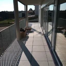 Nowoczesny balkon z szarymi płytkami podłogowymi, metalową balustradą rzucającą cienie i przeszklonymi drzwiami balkonowymi, widoczne kartony i narzędzia.