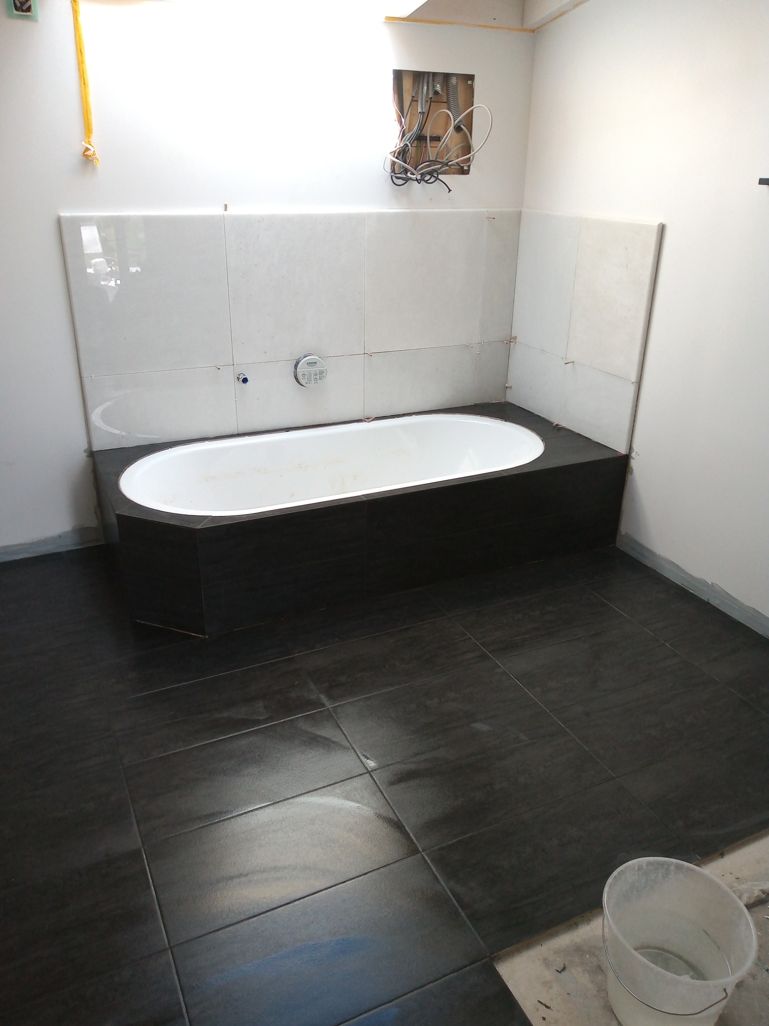 Wnętrze łazienki w trakcie prac wykończeniowych; widoczna wanna obudowana ciemnymi płytkami, białe płytki na ścianie, ciemna podłoga z płytek oraz odkryte przewody elektryczne w ścianie.