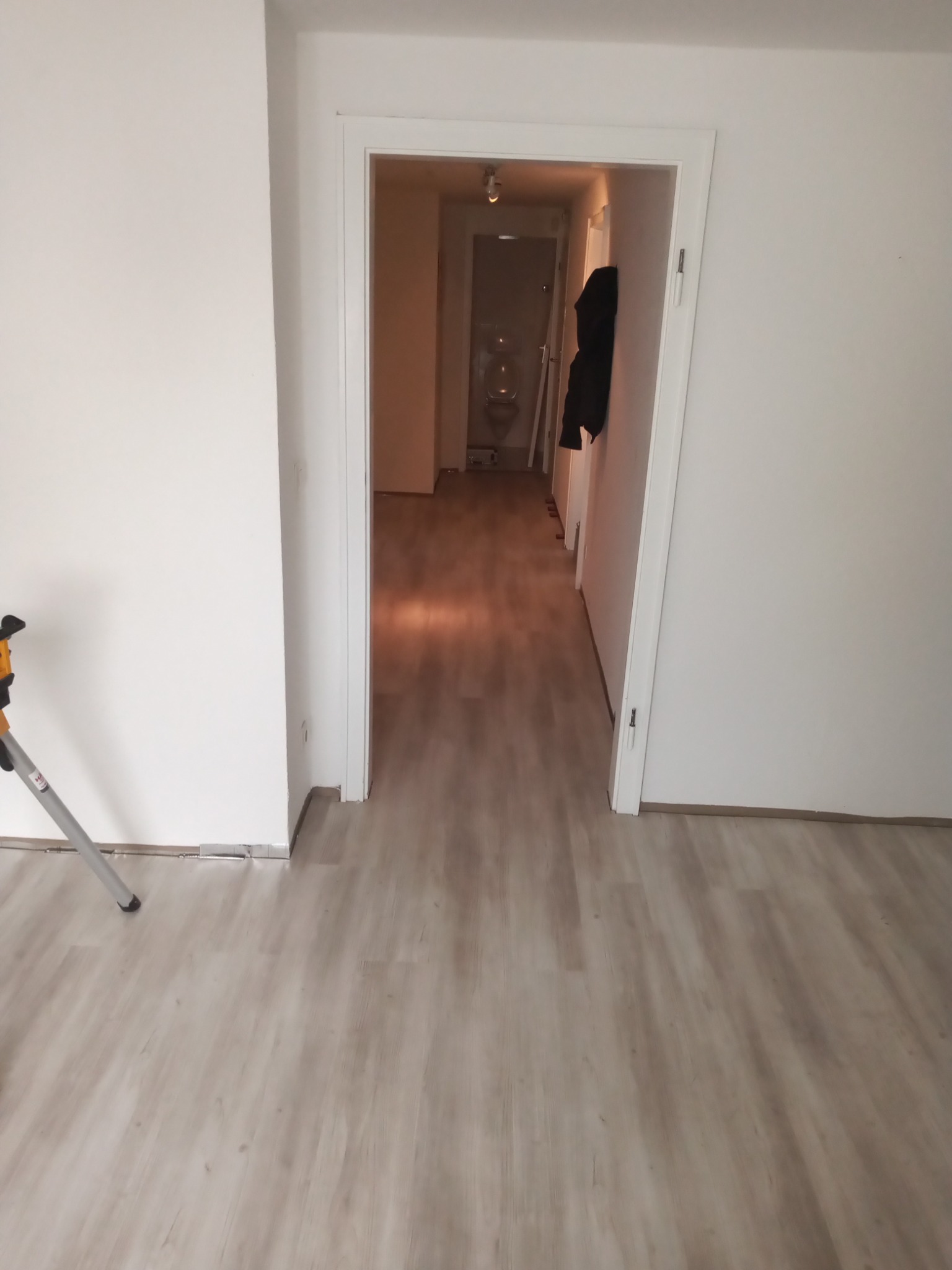 Nowo położona jasna wykładzina podłogowa w pokoju z białymi ścianami, widok na otwarte drzwi do łazienki, w rogu widoczny statyw.