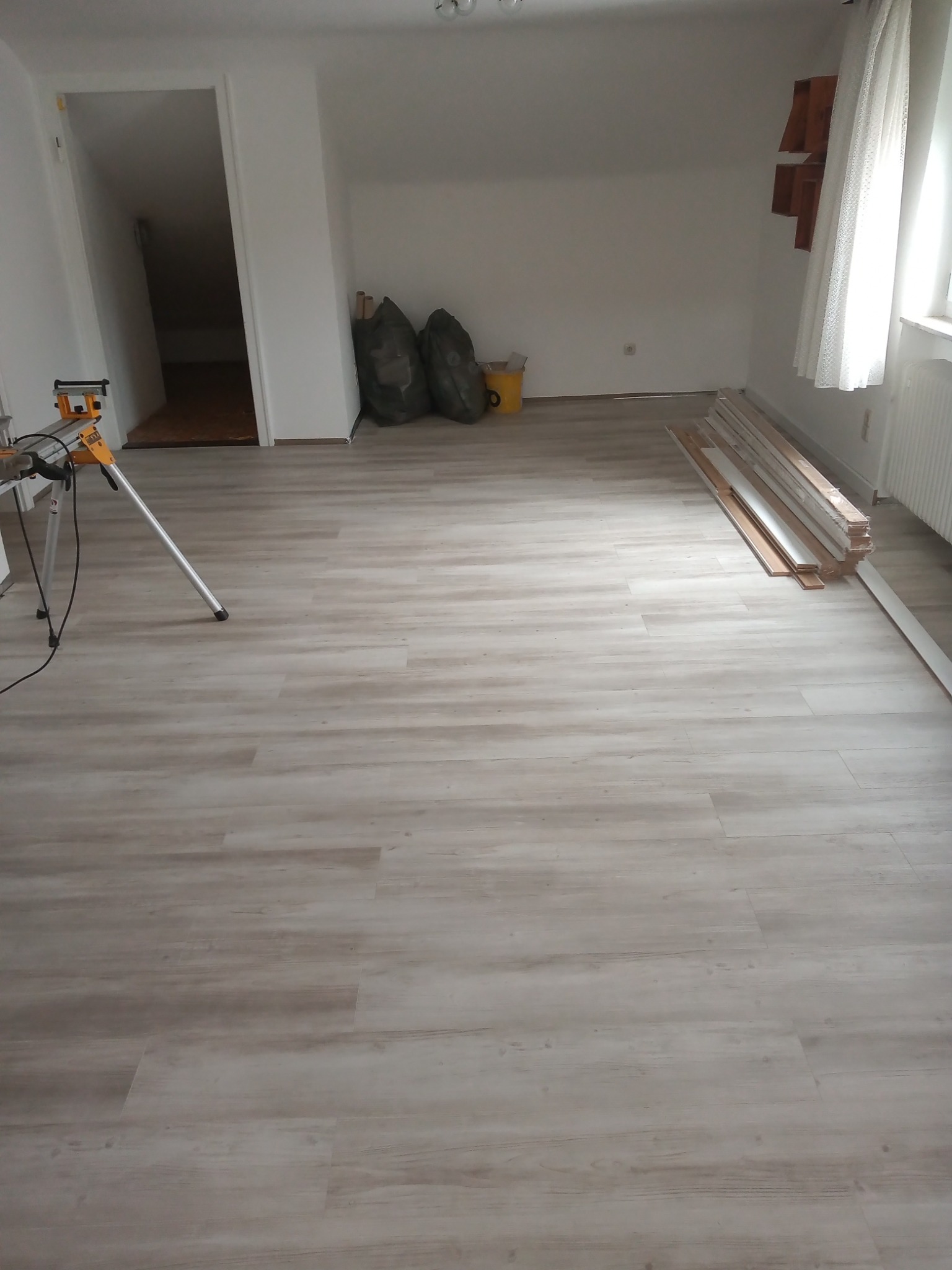 Nowo położona podłoga z paneli laminowanych w jasnym odcieniu drewna, widoczne narzędzia i materiały przygotowane do dalszego montażu, w tle worki i wejście do pomieszczenia gospodarczego.