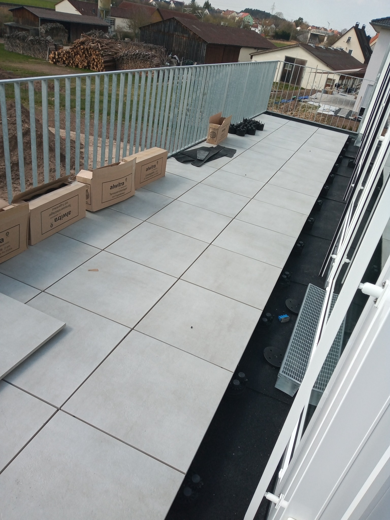 Montaż płytek na tarasie z widocznymi pudełkami po płytkach i elementami montażowymi, w tle zabudowa jednorodzinna i składowane drewno.